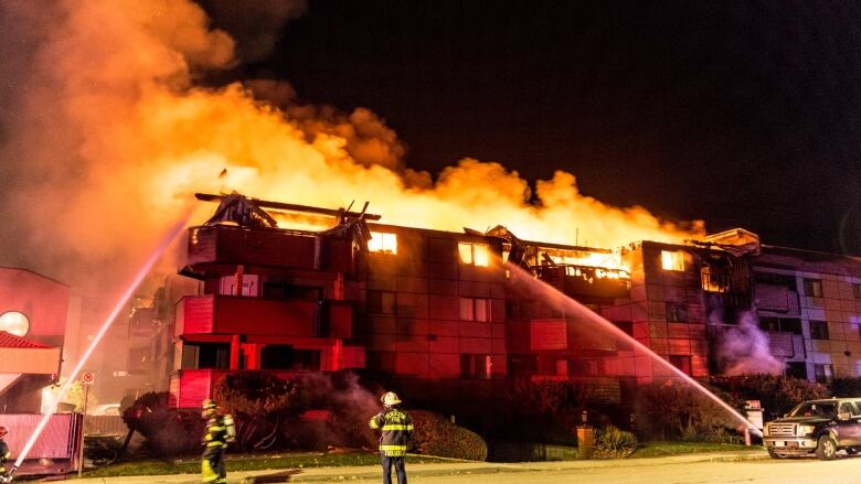 photo of a fire fighters using hoses to spray water on a three story apartment complex that is burning in the dark of nigh time. 