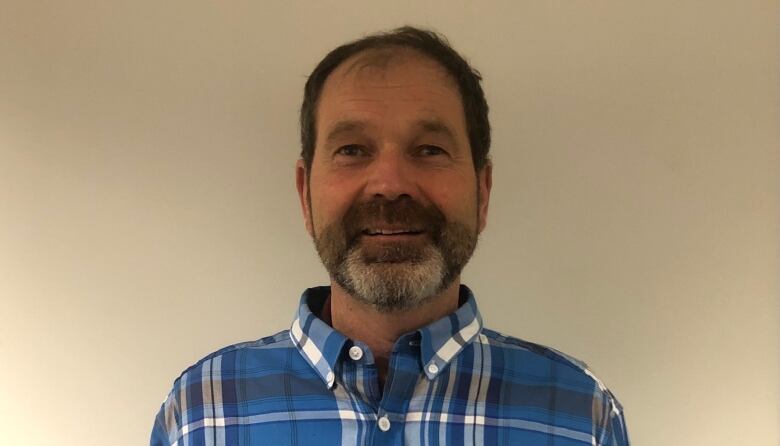 A bearded man in a plaid shirt stands in front of a blank wall