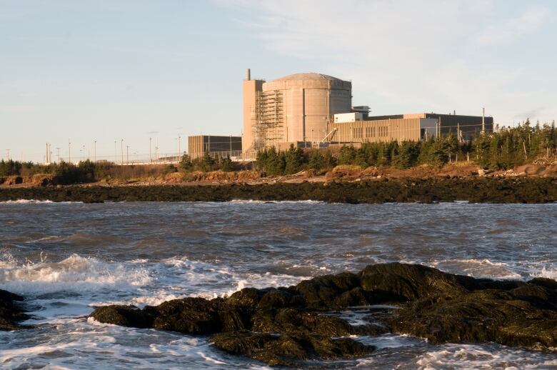 A concrete building with a squat, round tower sits on the edge of land with rocks and water in the foreground.