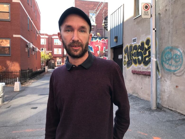 A man wearing a sweater and a cap stands in an alleyway.