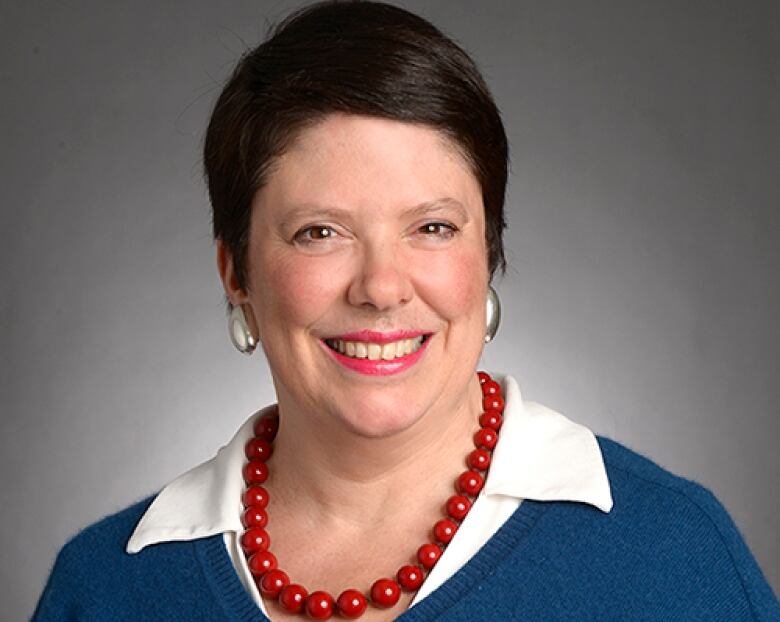 A woman with short brown hair is wearing a white blouse, blue sweater and a red necklace.