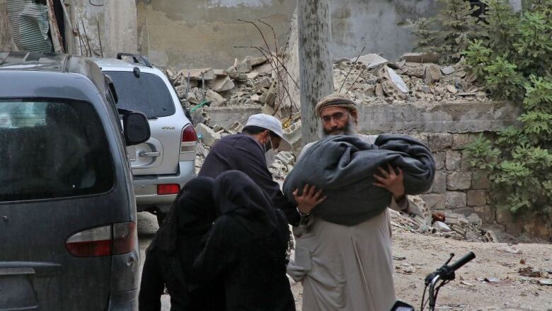 A man carries a body in a blanket past the rubbled of a destroyed building.