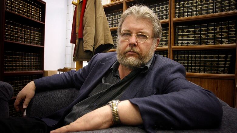 A bearded man with glasses sits in an office, in front of shelves lined with books.