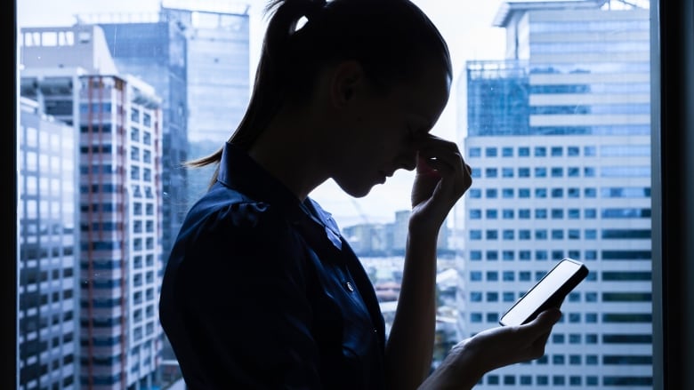 A woman is shown holding her nose and looking at her phone in front of a window. 