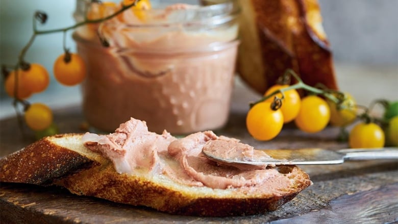 closeup on chicken liver mousse on a slice of baguette on a wooden cutting board. a silver knife is leaning on the slice, cherry tomatoes and a jar of the mousse are in the background. 