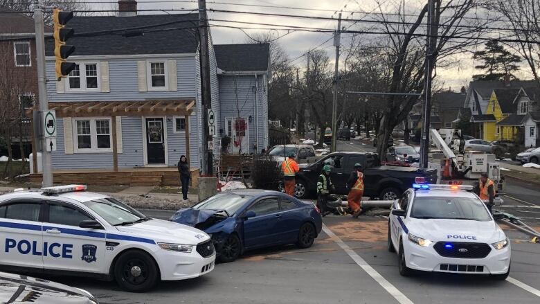 A blue car in the middle of an intersection has a crumpled front hood, while police cars sit surrounding it