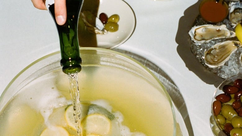 overhead shot of a hand pouring a bottle of champagne into a punch bowl with lemons and lemon leaves in it. the bowl is on a table with a white tablecloth and bowls of olives and oysters are in the right side. 