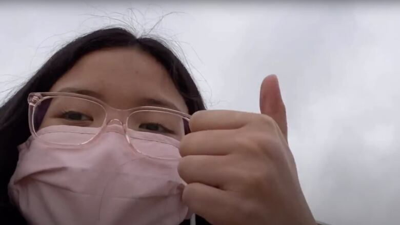 A teenaged girl with black hair and glasses, wearing a face mask, gives a thumbs up to the camera.