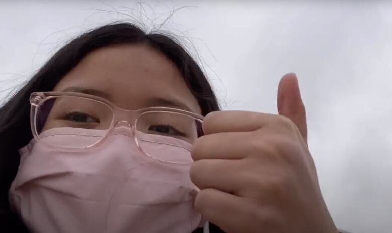 A teenaged girl with black hair and glasses, wearing a face mask, gives a thumbs up to the camera.