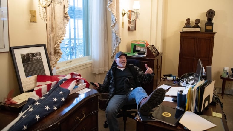 A man in blue jeans, a blue plaid jacket and a baseball cap is seen sitting at a desk, with one foot propped up on it.