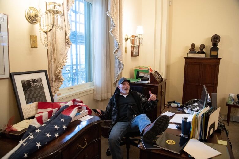 A man in blue jeans, a blue plaid jacket and a baseball cap is seen sitting at a desk, with one foot propped up on it.