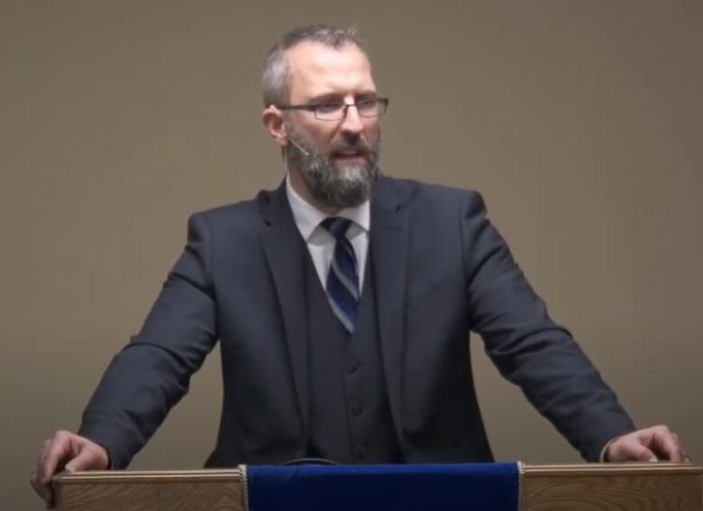 a man in a grey suit and beard speaks at a podium 