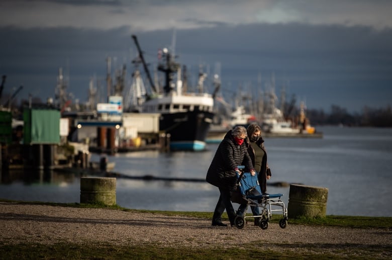 People wearing face masks to curb the spread of COVID-19 walk together at Garry Point Park as fishing vessels docked at Steveston Harbour are seen in the distance, in Richmond, B.C., on Sunday, Jan. 10, 2021.