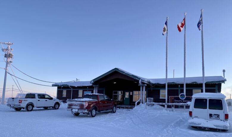 The community government office in Wekwet in winter. A one-story building with three flag poles and trucks parked out front.