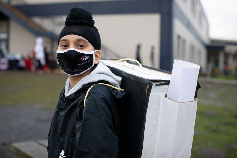 A Sikh boy wearing a black coat and black facemask carries a backpack.