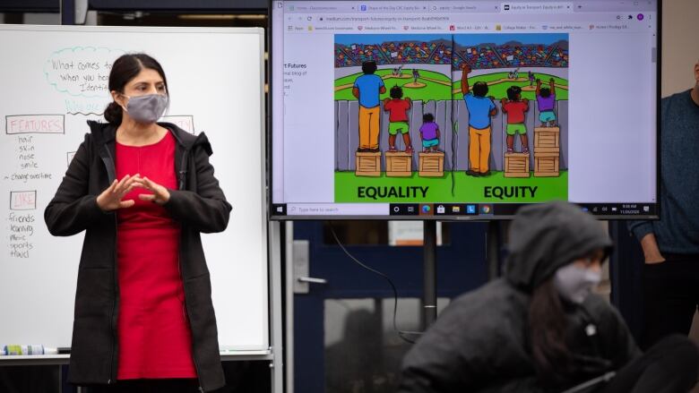 A female teacher stands in front of a whiteboard wearing a black coat and grey facemask.