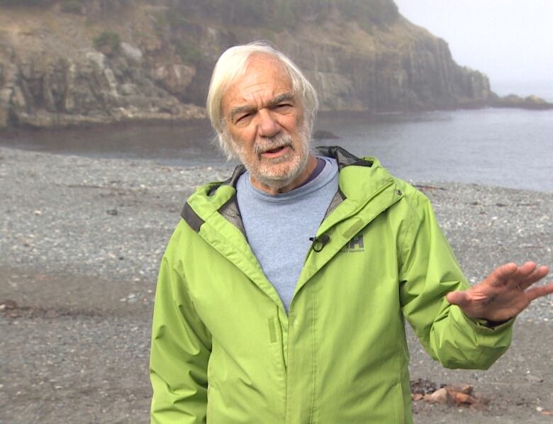 A man in a windbreaker jacket poses on a beach in front of the ocean.