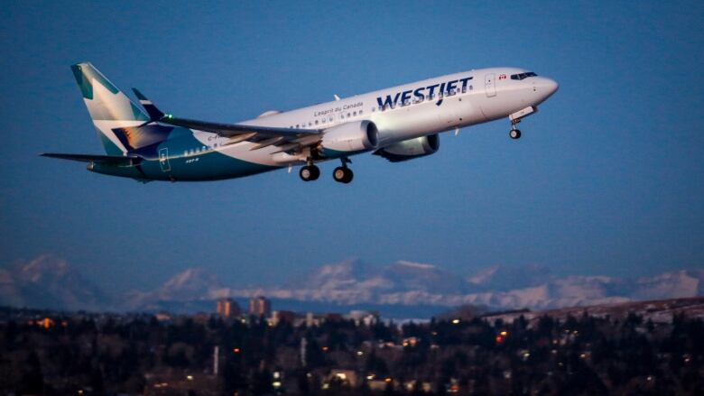 A jetliner branded WestJet takes off against a blue sky  and mountains in the distance.