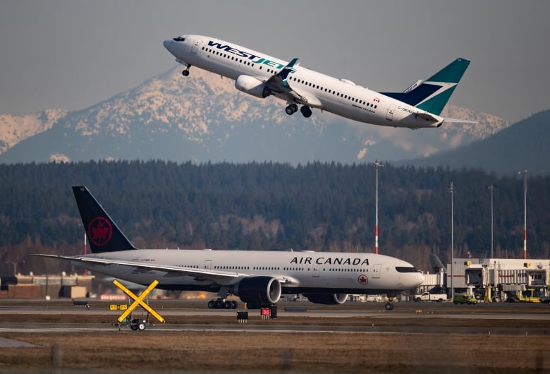 An Air Canada plane is driving on a runway underneath a WestJet plane that is taking off in front of a mountain range.