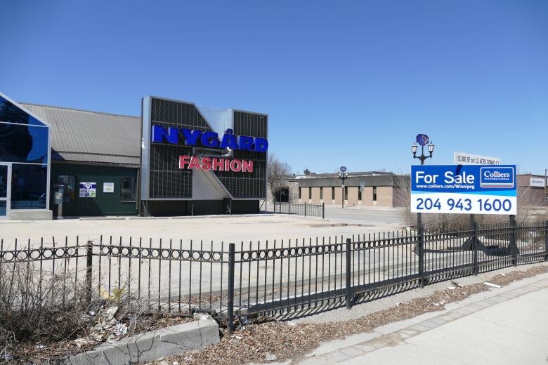 The photo shows a Nygard Fashion store on Broadway in Winnipeg. The building is surrounded by a black metal fence and there are no cars in the parking lot. There is a for sale sign in front of the building.