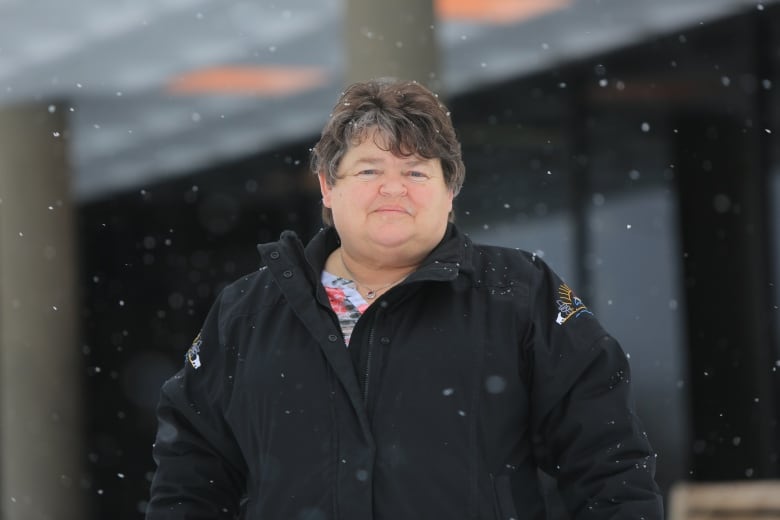 A woman in a black jacket poses for a photo as snow falls down.