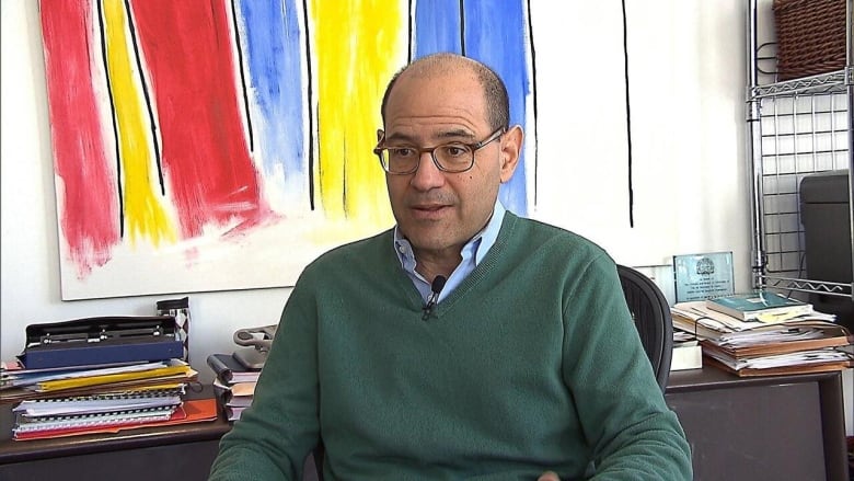 A man wearing a sweater over a collared shirt sits in an office with bright-coloured abstract art hanging over the desk behind him.