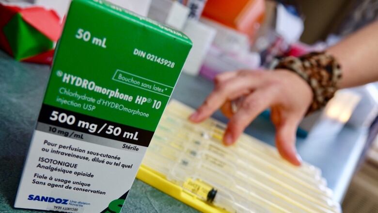 A green box of medication is pictured. A person's hand is visible in the background with  a tray of medical supplies.