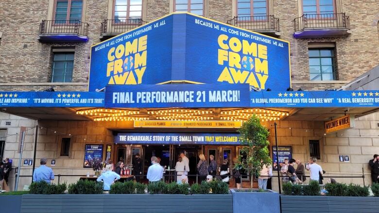 A dramatic sign displays the words 'Come From Away' above a theatre's doors. A small lineup of about 15 people are waiting outside.