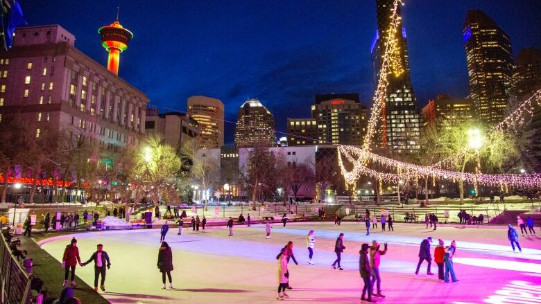 Skaters enjoy an outdoor ice rink under the lights at night surrounded by downtown towers.