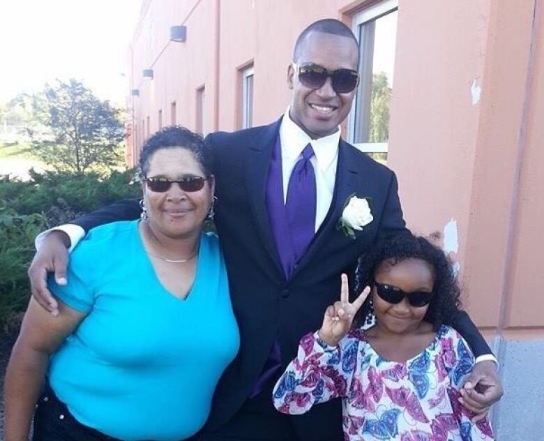 A tall Black man in a suit with his arms around a smaller older woman wearing a blue shirt and a girl making a peace sign. All are smiling and wearing sunglasses.