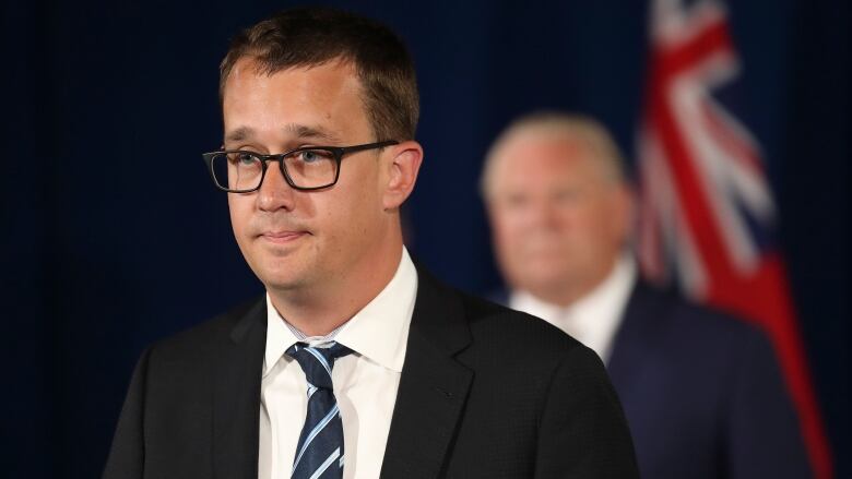 A man speaks at a press conference. Another man is seen in the background.