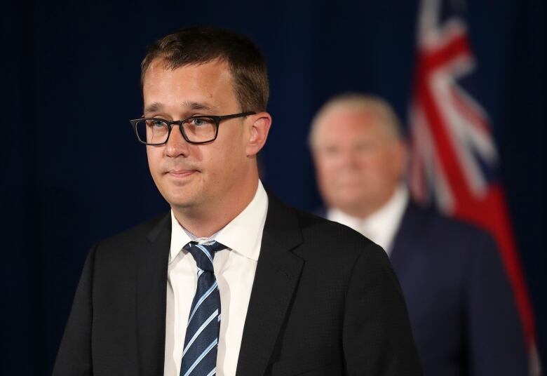 A man speaks at a press conference. Another man is seen in the background.