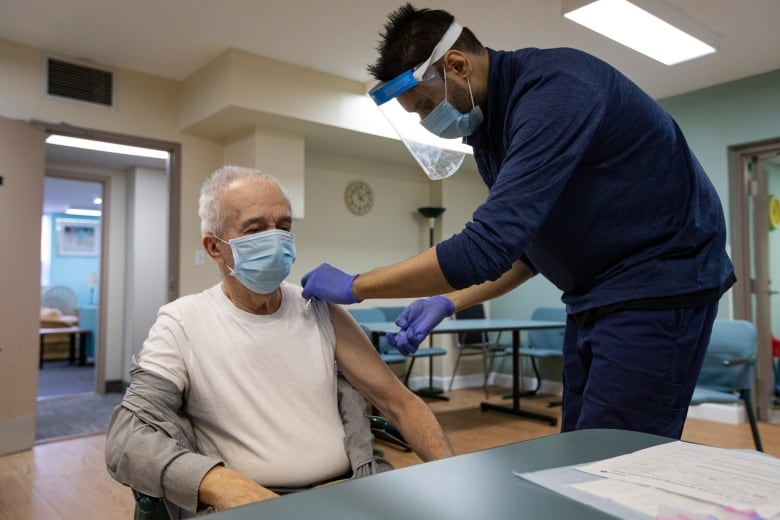 A medical worker wearing a face shield and mask prepares to give a vaccine to a seated 77-year-old man.  