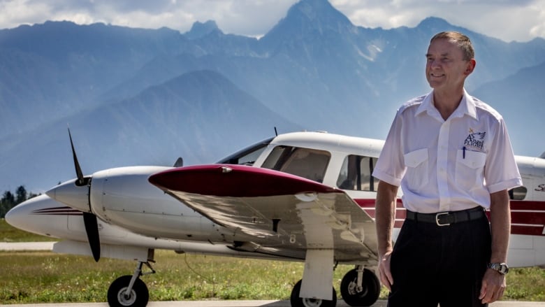 A man stands in front of a plane with mountains in the background.