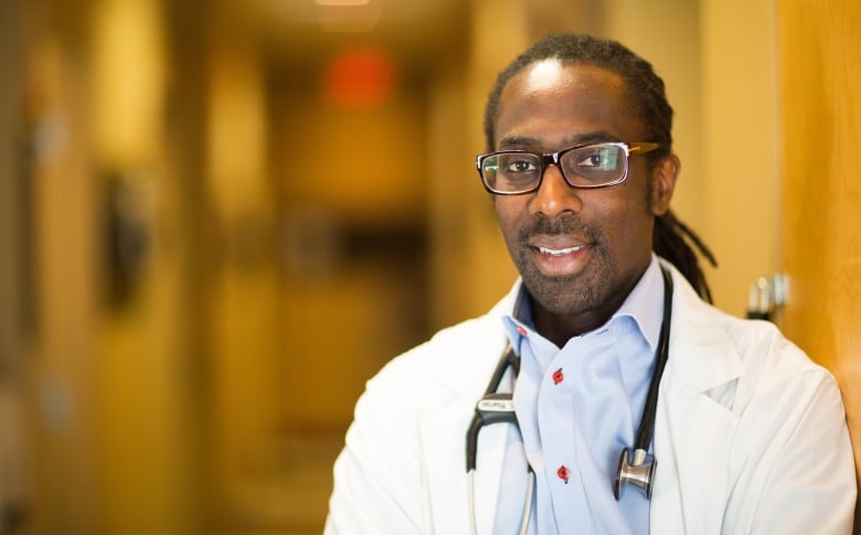 A man with glasses and a white coat stands in a hallway. 