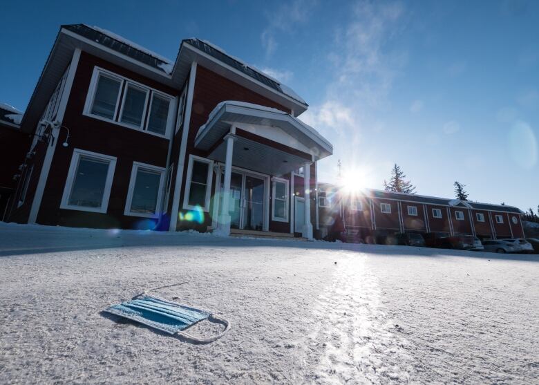 A red building with several rooms stands with the sun shining in the background, a blue mask sits on the frozen, snowy ground in front.