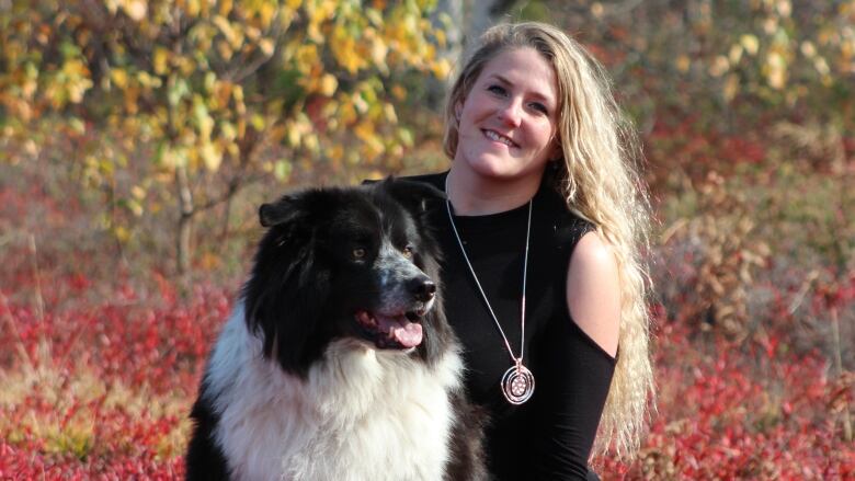 A smiling young woman with long blond hair and a large, black and white dog, surrounded by brilliant-red coloured folliage of a blueberry field. 