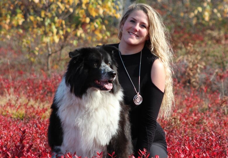 A smiling young woman with long blond hair and a large, black and white dog, surrounded by brilliant-red coloured folliage of a blueberry field. 