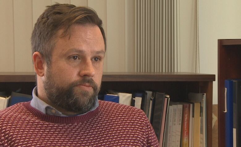 A man in a sweater sits in front of shelves filled with binders and books.