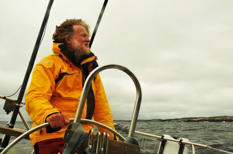 A man with grey hair wearing a raincoat steering a sailboat.