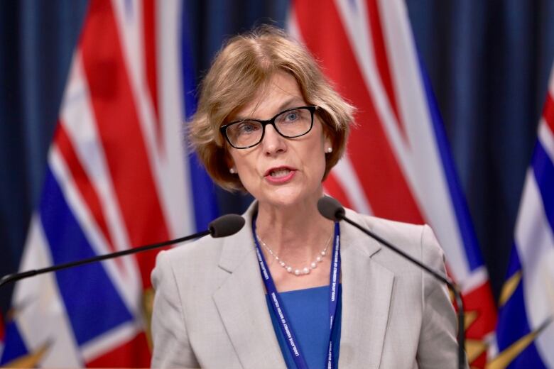 A white woman wearing a grey blazer speaks in front of two B.C. flags.