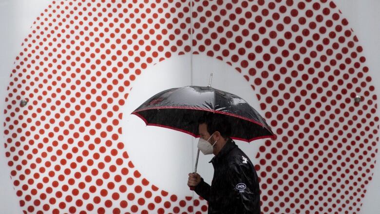 A man wears a mask and carries an umbrella in front of a circular red-and-white logo.