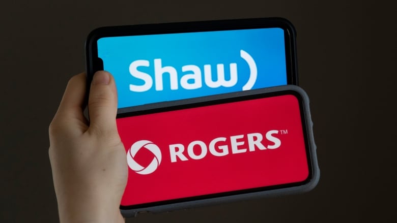 Close-up of a hand holding two cellphones that clearly display the Rogers and Shaw Communications names and logos. 