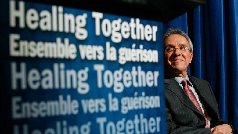 A man with grey hair and glasses sits next to a sign that says 'healing together' in French and English. 