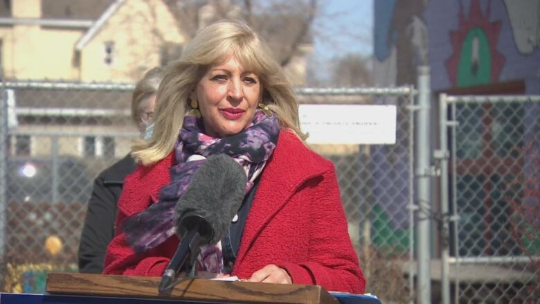 A woman in a red coat speaks at a podium outside.