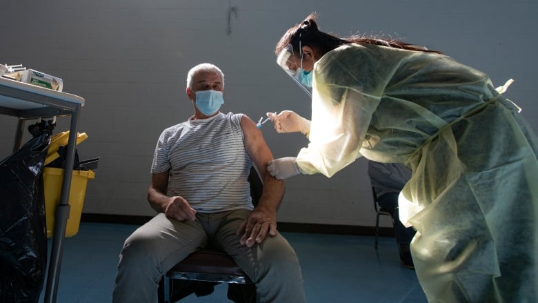 A health-care worker wearing a yellow gown over her clothing, along with a face shield and face mask, administers a vaccine into the arm of an elderly man.