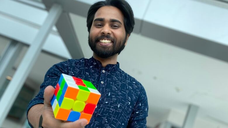 A man smiles at the camera while holding out a Rubik's cube in one hand. 