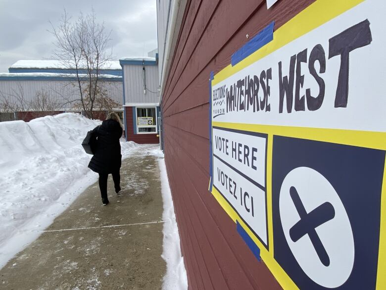 A person is seen from the back walking toward the door of a school building, with a 'Vote Here' sign in the foreground.