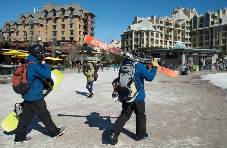 Two people carrying skis and snowboards walk across a brick pathway