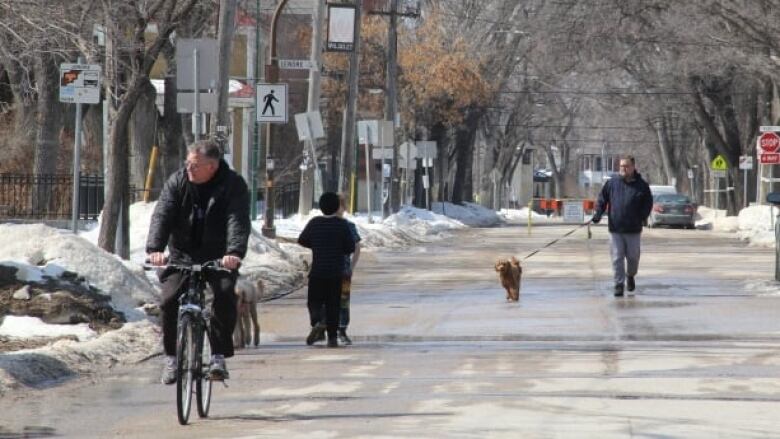 A man bikes down a closed-off street while another man walks his dog. 